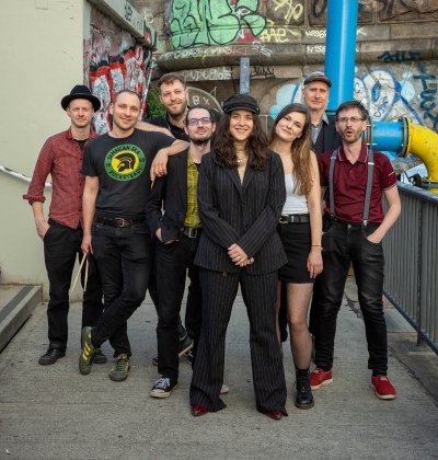 Foto der Band Arte Bella & The Fellas. Eine Gruppe von acht Personen steht auf einem urbanen Gehweg mit Graffiti-Wänden im Hintergrund. Alle Personen schauen freundlich in die Kamera. Die Gruppe ist gemischt, mit Männern und Frauen, alle in lockerer, indi